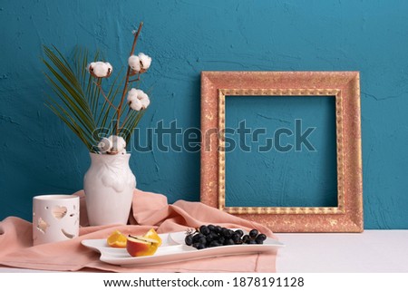 picture frame, vase with dry cotton plant and breakfast, grapes and oranges on a blue background on the table
