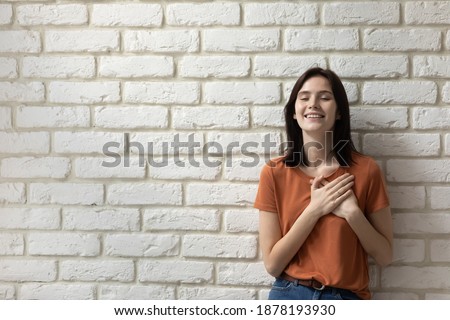 What a relief. Happy young woman posing close to wall of bricks with closed eyes feeling grateful hopeful. Millennial female putting hands on chest over heart express sincere feelings. Copy space