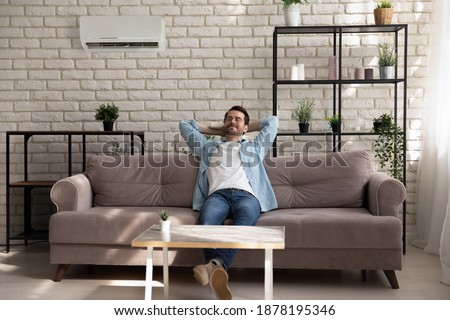 Paradise. Serene young guy with closed eyes reclining on couch at modern designed living room. Carefree millennial male enjoy breathing cool fresh air turn conditioner on using remote control device Royalty-Free Stock Photo #1878195346