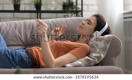 Music lover. Carefree young female in wireless headset resting on sofa lying with closed eyes relieving stress. Calm lady listen to playlist of favorite songs snap fingers feel rhythm enjoy melody Royalty-Free Stock Photo #1878195634