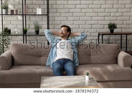 Lazy morning. Successful millennial man relaxing on comfy sofa at home after finishing work looking aside with glad smile. Calm young guy taking pleasure in awaited weekend enjoying peaceful dreams Royalty-Free Stock Photo #1878196372