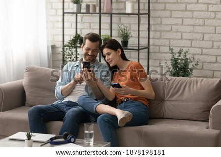 Sharing information. Happy loving young spouses sitting on couch at living room using smartphones exchanging files. Couple of diverse friends showing each other funny photo video at social network