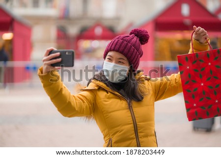 Asian girl enjoying Christmas shopping during covid19 - young happy and beautiful Chinese woman holding red shopping bag taking selfie with presents on xmas street market
