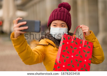 Asian girl enjoying Christmas shopping during covid19 - young happy and beautiful Chinese woman holding red shopping bag taking selfie with presents on xmas street market