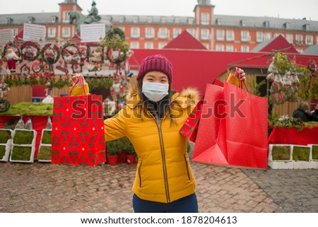Asian girl enjoying Christmas shopping during covid19 - young happy and beautiful Chinese woman with mask holding red shopping bag buying presents on xmas street market