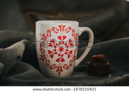 White ceramic coffee cup with a red snowflake graphics on a gray natural fabric. Winter cozy background with chocolate decoration.