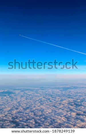 Trail from an airplane high in the blue sky above the clouds. View of a flying plane from another plane, below the clouds. Porthole view of clouds and blue sky. Cumulus clouds top view.