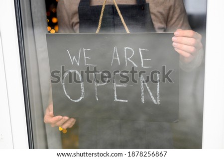 Cafe barista in apron is holding open sign board in hands, view through glass door, a face is not visible
