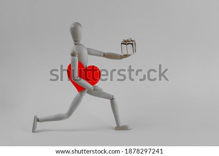 A black and white little wooden man carries a red heart and a gift on a gray background under his arm. Valentine's Day background.