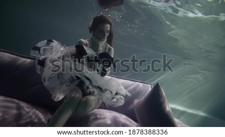 A female model in a long dress and heels poses beautifully on a gray sofa with pillows, which is under water. bubbles float around. Her skirt and hair flutter under the water.