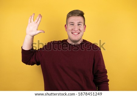 Young blonde man wearing a casual red sweater over yellow background doing hand symbol
