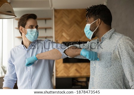 Smiling young multiracial male colleagues in facial mask and rubber gloves greeting touching elbows. Smiling diverse men make gesture say hello during coronavirus pandemics. Covid-19, corona concept. Royalty-Free Stock Photo #1878473545