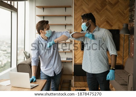 Smiling African American man greet with Caucasian colleague in office on covid-19 quarantine. Happy multiracial male employees in facial masks and rubber gloves touch elbows. Corona concept. Royalty-Free Stock Photo #1878475270