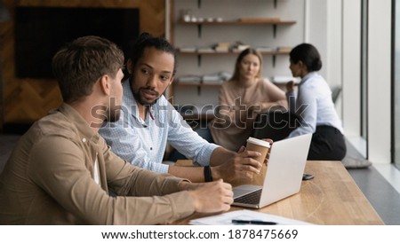 Wide banner view of multiracial employee sit at desk in office work on laptop together discuss project. Focused young diverse businesspeople cooperate on computer brainstorm at meeting in office.