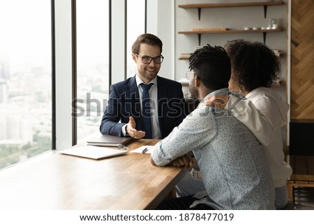 Smiling Caucasian male realtor talk with biracial couple at meeting, consult about house purchase or rent. Real estate agent have consultation with African American family clients about home rental. Royalty-Free Stock Photo #1878477019