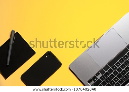 Flat lay composition with a silver grey laptop, black smartphone, notepad and a pen on an isolated yellow background.