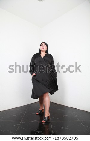 Ready to go. Overweight and carefree. Woman in oversize black silk dress posing on white wall background in studio. Stylish oversize clothes for plus size women. Modern life. Fashion shop. Diversity
