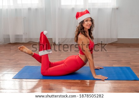 A beautiful sportive girl is engaged in yoga, pilates, exercises, body training. A woman in a red sports uniform against the background of the training hall. Santa hat on the head. Healthy lifestyle