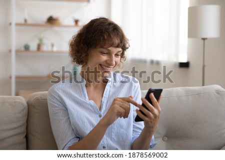 Happy young female rest on sofa at living room making video call by cell shooting selfie using phone webcam. Smiling woman enjoy chatting in app scrolling web pages reading electronic book good news