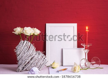 burning candle, writing materials, roses in a vase, white photo frames, wooden heart on a red background on the table