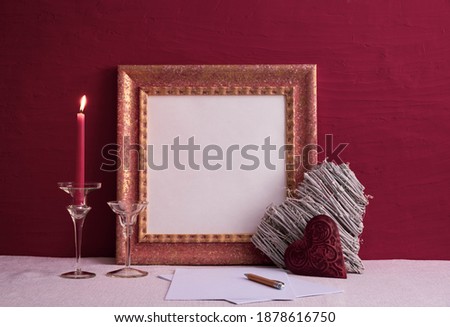 a candlestick on a long leg with a burning candle, two decorations in the shape of a heart, an old photo frame on a red background on the table
