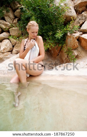 Attractive young woman relaxing by the edge of a natural transparent swimming pool in a health spa enjoying a summer holiday trip against a wall of rocks and stones. Healthy travel lifestyle outdoors.