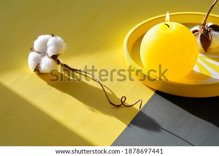 Illuminating Yellow and Ultimate Gray, colors of the year 2021. Burning candle on decorative tray and cotton flower on split yellow grey paper background. Sunlight, long shadows, isometric projection