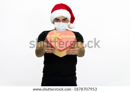 Young man wearing a Santa hat. She is holding a heart-shaped box in her hand. There is a medical mask on the music. He is wearing a black shirt. Isolated image white background.