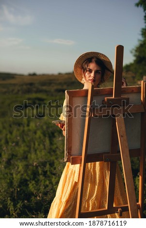 A girl artist in a yellow long dress and a straw droplet draws a picture on an easel in the field.