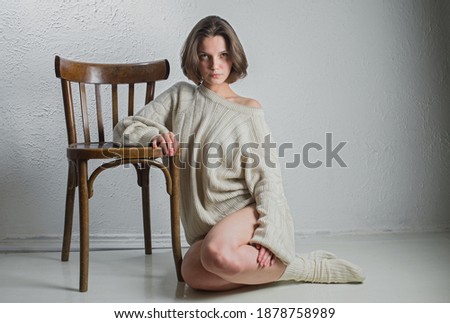 A young and beautiful girl in a warm knitted sweater sits near a wooden chair. Light background.