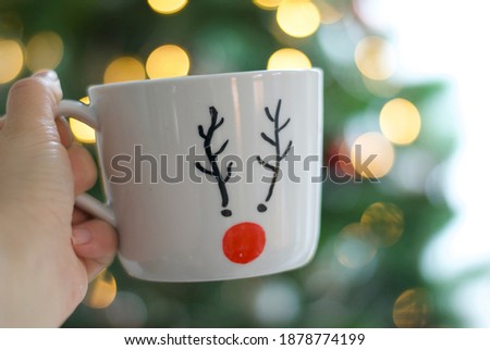 A hand holding a white mug with a picture of a  reindeer, a green Christmas tree is on the background.