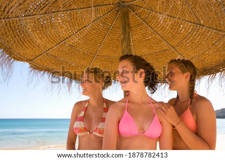 Close-up shot of young girls looking away in a swimming suit posing under a beach umbrella on a sunny day with the ocean in the background.
