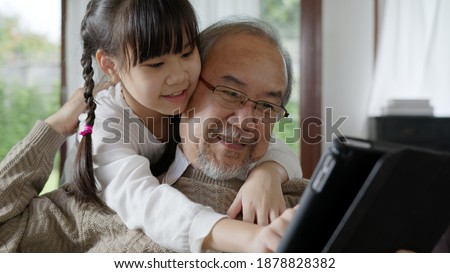 Candid of old senior asian grandparent play and watch with kid grandchildren with technology on computer tablet at home in bonding relationship in family. Young girl hug older man from back. Royalty-Free Stock Photo #1878828382