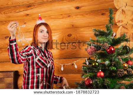 A cheerful woman in a plaid shirt hangs a garland on a Christmas tree oon the background of a lighted fireplace, hanging socks for gifts. Christmas and new year home preparation