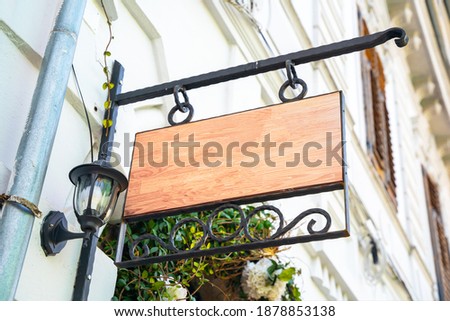 Empty wooden sign with wrought-iron frame and vintage street lamp hangs on the wall of house outside. Blank for advertising or signage of store, cafe, or public organization