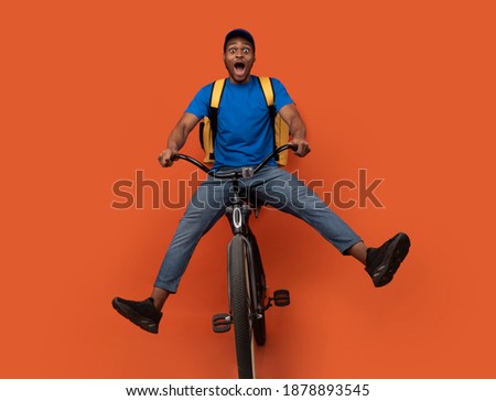 Full length of funny black delivery man in blue cap and uniform riding bicycle, wearing yellow thermo backpack bag isolated over bright orange blue studio background. Fast and quick shipment service