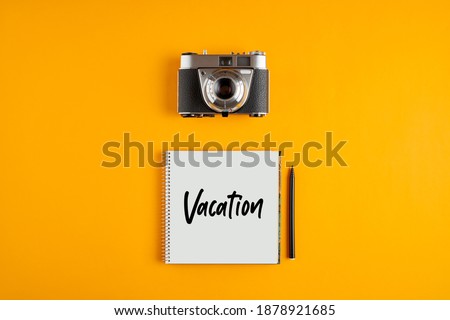 Vintage camera with a notebook with the word vacation against yellow background. Vacation photography concept.