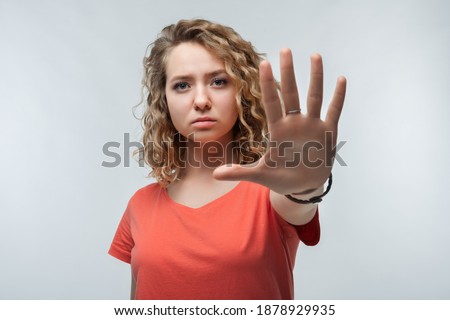 Image of serious blonde girl with curly hair in casual t shirt doing stop sign with palm of the hand. Warning expression on the face. Studio shot, white background. Human emotions concept