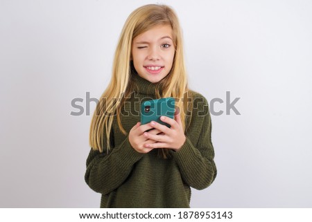 Cute Caucasian kid girl wearing green knitted sweater against white wall taking a selfie  celebrating success