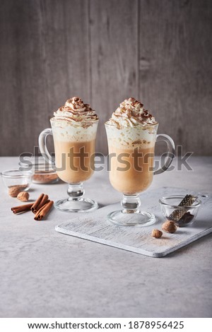 Two pumpkin spice lattes in tall latte glasses, topped with whipped cream and chocolate dust, on light grey table. Near them: ground spices in glass bowls, whole nutmegs, small grater, cinnamon sticks