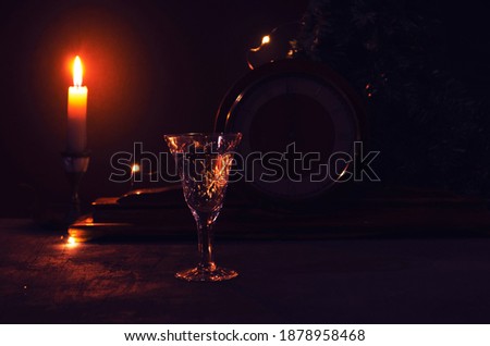 Glass with white wine with vintage old wooden clock and burning yellow candle  garland lights on the background. Midnight on clock. Night festive picture for background. Beautiful golden lights.