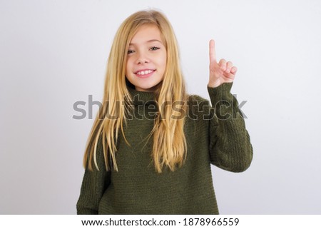 Cute Caucasian kid girl wearing green knitted sweater against white wall showing and pointing up with finger number one while smiling confident and happy.