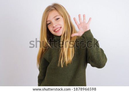 Cute Caucasian kid girl wearing green knitted sweater against white wall Waiving saying hello happy and smiling, friendly welcome gesture.