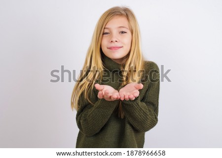 Cute Caucasian kid girl wearing green knitted sweater against white wall holding something with open palms, offering to the camera.