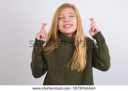 Cute Caucasian kid girl wearing green knitted sweater against white wall has big hope, crosses fingers, believes in good fortune, smiles broadly. People and wish concept