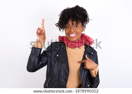 Young beautiful African American woman wearing knitted sweater against white wall says: wow how exciting it is, has amazed expression, indicates something. One hand on her chest and pointing with othe