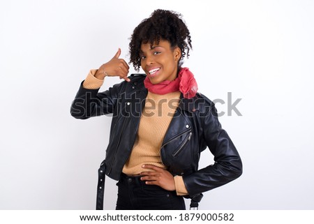 Young beautiful African American woman wearing biker jacket against white wall imitates telephone conversation, makes phone call gesture with hands, has confident expression. Call me!