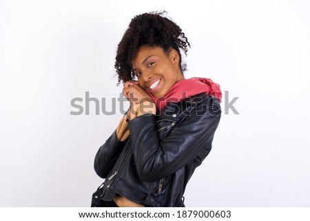 Dreamy Young beautiful African American woman wearing biker jacket against white wall, with pleasant expression, closes eyes, keeps hands crossed near face, thinks about something pleasant