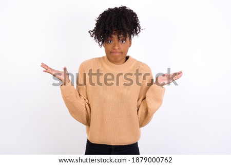 Puzzled and clueless Young beautiful African American woman wearing knitted sweater against white wall, with arms out, shrugging shoulders, saying: who cares, so what, I don't know. Negative human emo