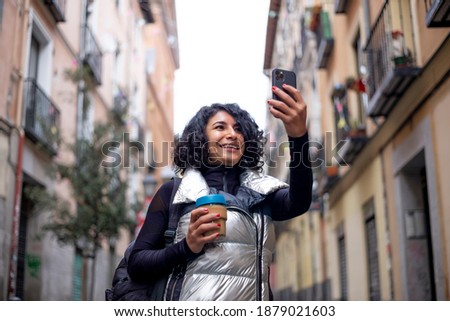 Curly black hair woman with coffee in hand using mobile phone in city with silver jacket